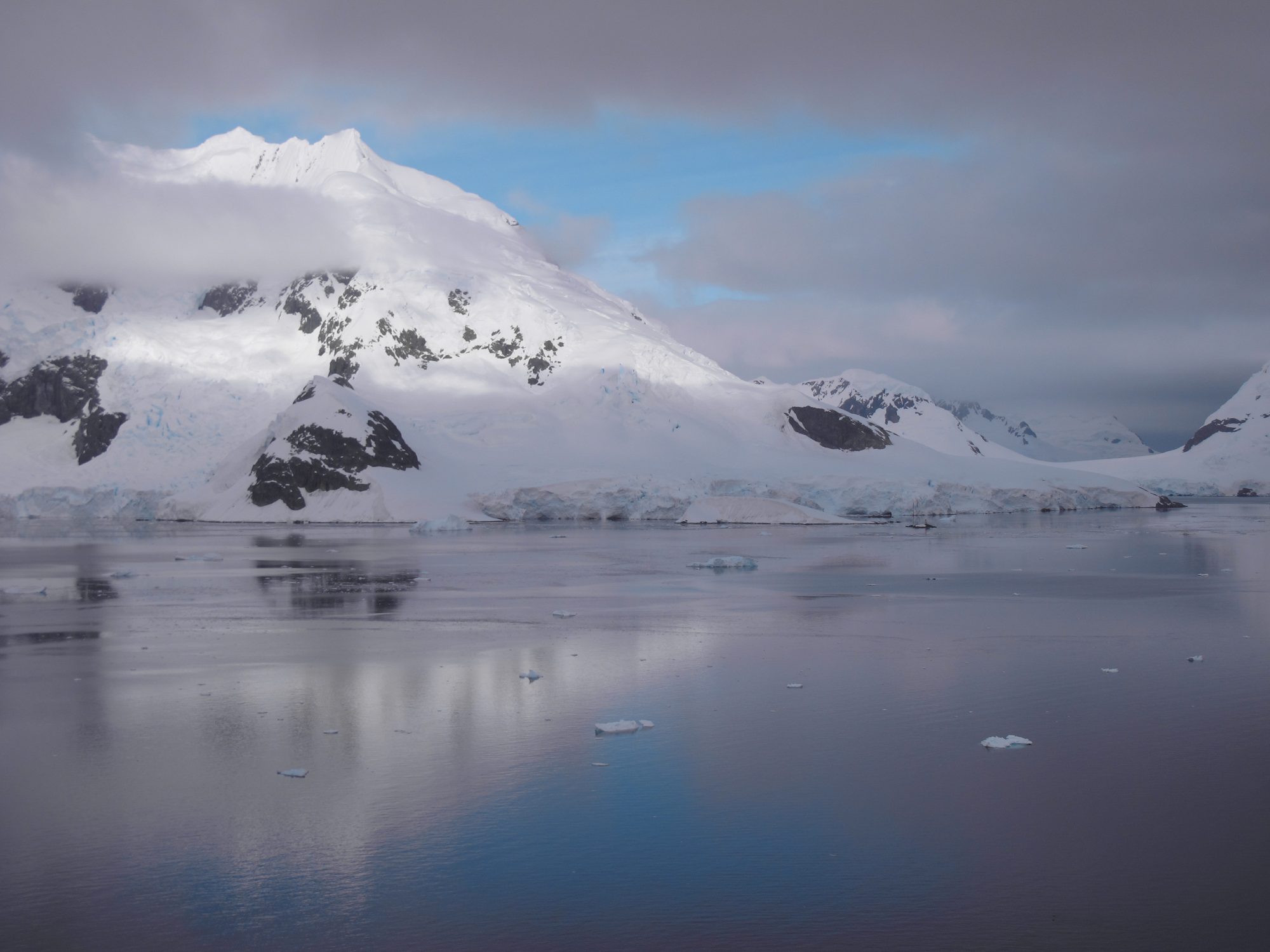 Cloud sitting on a snow peak in cool grey and blue, above calm water.