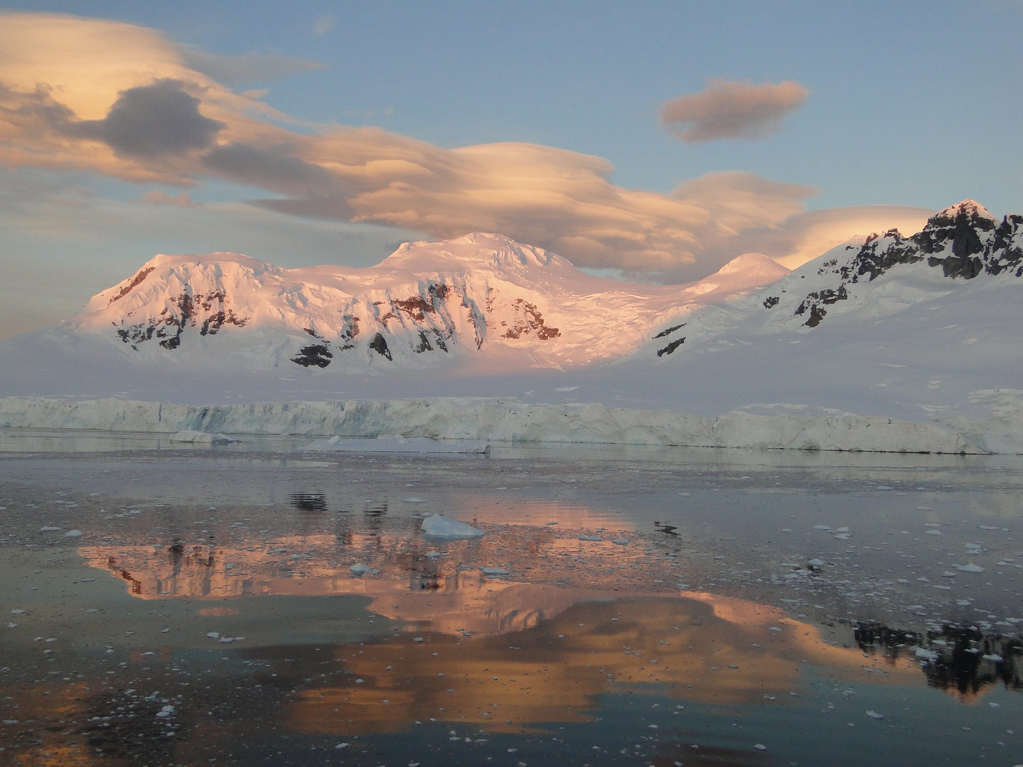 Alpenglow pink on a range above an ice cliff, reflected in still water.